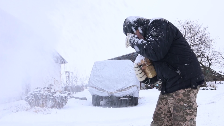 A man carries firewood during a heavy snowstorm. He shields his face from the snow.