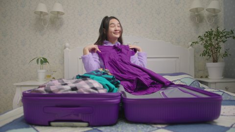 Happy Asian woman packing suitcase for vacation sitting in bedroom alone. 20s female dreaming of upcoming holidays holding color dresses.