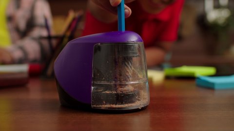 Close-up of child hand sticking pencil into electric pencil sharpener and sharpening while African American kids drawing picture in domestic interior.