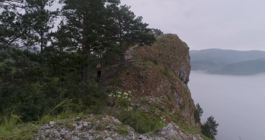 Girl traveler with backpack walks along ridge of rock in forest among fog. High quality 4k footage