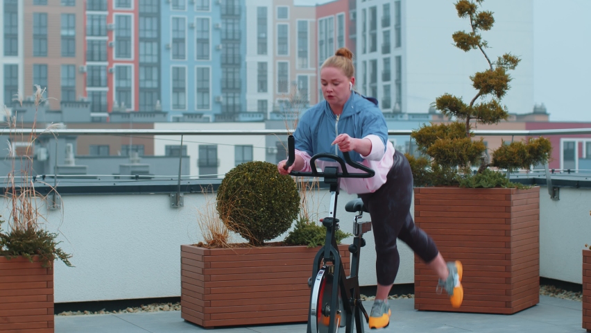 Caucasian woman coach spinning on stationary bike on house roof. Athletic girl performing aerobic riding training exercises on cycling bike. Weight Loss Cardio Healthy Lifestyle. Modern sport outdoors