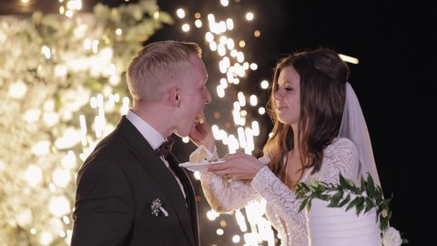 Newlyweds cut the wedding cake with knife. Fireworks. Lovely bride and groom couple eating wedding cake dessert. Lovely man woman make the first cut of three-tiered wedding cake. Outdoors slow motion