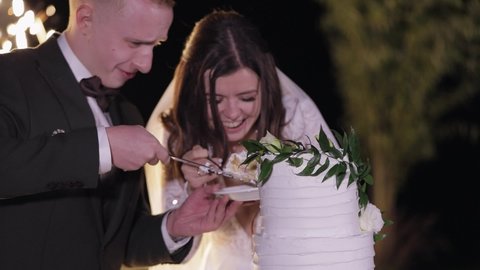 Newlyweds cut the wedding cake. Fireworks. Lovely bride and groom couple cutting wedding cake dessert with knife. Lovely man woman make the first cut of three-tiered wedding cake. Outdoors slow motion