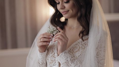 Close-up of beautiful lovely stylish smiling bride girl looking at gypsophila flowers bouquet at home. Wedding morning preparations before ceremony. Bride woman face with fashion hairstyle make-up
