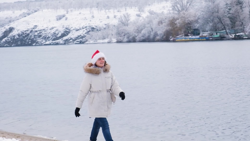 A woman in a Santa Claus hat walks along the bank along the river in winter and smiles, there is snow