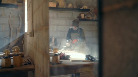 Reflection in mirror of female carpenter in protective uniform using angle grinder for polishing piece of wood at workbench in carpentry on Mallorca
