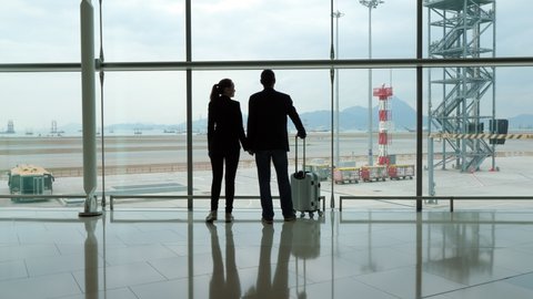 Passengers stand against terminal window, woman and man look to airfield. Lady turn head and speak to partner, he answer. Silhouetted shot at empty hall, no movement outside