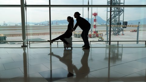 Hilarious two entertain at empty airport terminal, woman ride wheeled bag, man push her forward. Travelers couple have fun after long flight or spend time before departure