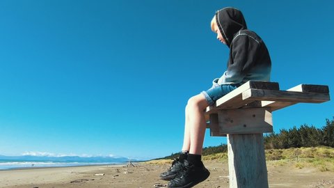 Slow Motion Zooming Out View Of Boy Sitting On A Pole On Woodend Beach, Pegasus Bay, Watching The Sea And The Waves - Dolly Shot