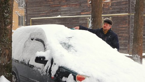 A man cleans the roof of a car from snow with a brush. Cloudy cool winter day, fresh snow is falling from the sky. Auto like a snowdrift. Climate change, city, snowstorm. UHD 4K.