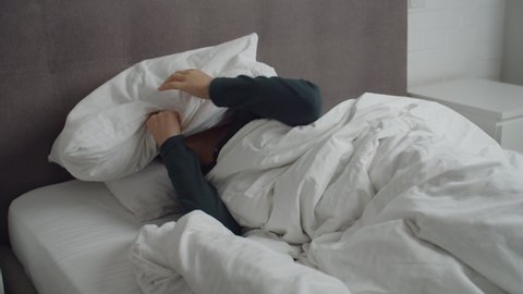 Sleepless adult female lying in bed, covering ears with pillow, can't sleep at home because of too loud sound, noisy neighbors. Irritated woman feeling uncomfortable, trying to relax indoors