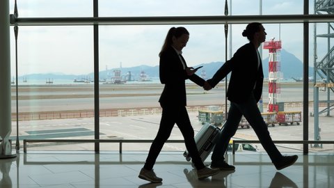 Man walk ahead, lead woman by hand, silhouetted shot at arrival area of international airport. Two people silhouettes pass by against glass wall, apron and empty taxiways seen outdoors