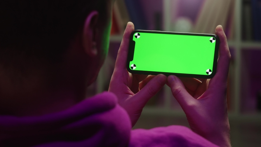 Man using smartphone with chroma key in evening. Young guy sitting on sofa and holding mobile phone with green screen, back view. Royalty-Free Stock Footage #1084609477