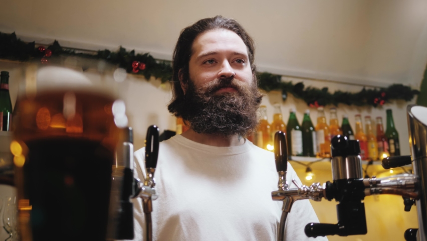 Beer barman portrait. Bearded man standing behind bar counter and smiling. Happy brewer close-up. Professional bartender posing in restaurant. 