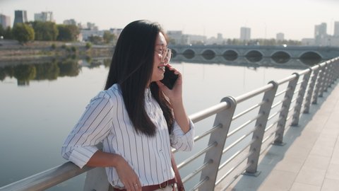 Medium shot of young beautiful Asian woman talking on smartphone standing at river embankment on sunny day