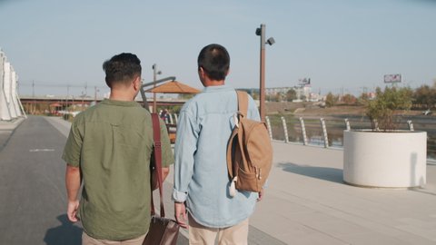 Tracking back-view medium shot of two young Asian men in casualwear walking outdoors in summer along embankment