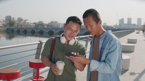 Medium shot of two young Asian male friends talking while scrolling on smartphone standing at river embankment on sunny day
