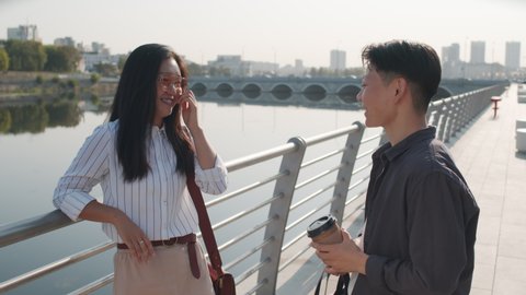 Medium shot of two young Asian female friends standing at embankment on sunny day and chatting