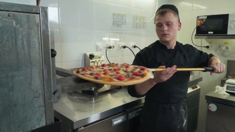 Cook Takes Prepared Delicious Italian Pizza From Oven At Kitchen. Cook Using Pizza Peel To Place Delicious Italian Dish In Box. Deliciously Cooked Italian Pizza With Meat And Tomatoes. Restaurant