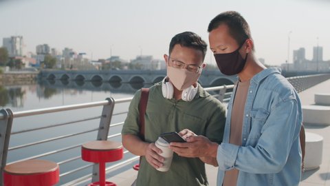 Medium shot of two young Asian men wearing face masks using smartphone and having conversation standing at river embankment on summer day