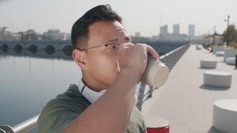 Close up portrait of young Asian man in eyeglasses drinking takeaway coffee and looking away standing at river embankment on sunny day