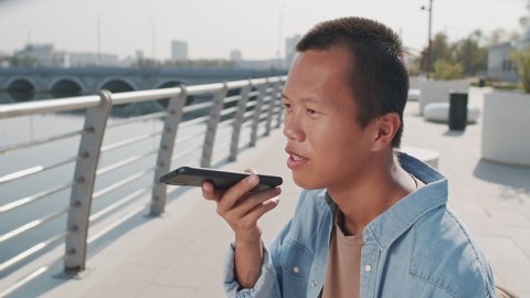 Medium closeup of young Asian man recording voice message on cellphone sitting at river embankment on summer day