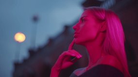Fashion model stand on dark street in neon pink lights, looking to camera. Portrait of young beautiful blonde woman in city lights. Close-Up Portrait. Side view