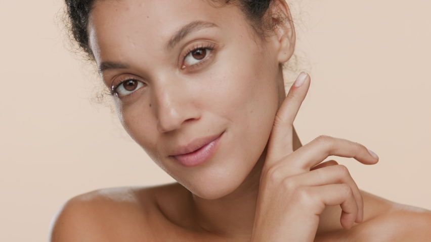 Good-looking dark-haired African American young slim woman with naked shoulders strokes her jaw line and smiles wide for the camera on beige background | Sagging jowls prevention concept