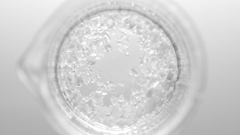 Top view macro shot of crystals fall down from test tube into clear fluid in flask creating different sized bursting bubbles on light grey background. Mineral skincare cosmetics formulating concept