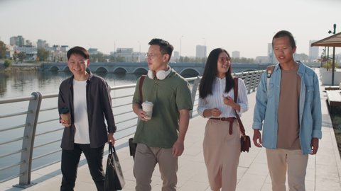 Tracking shot of four young male and female Asian friends walking along river embankment on sunny day and having conversation