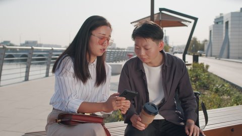 Medium shot of two young Asian female friends looking at smartphone screen having discussion, sitting on bench at river embankment on sunny day