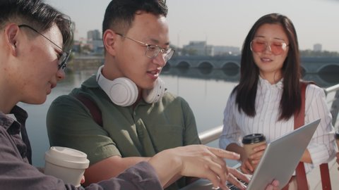 Medium shot of four young Asian colleagues coworking outdoors in summer, standing at river embankment with laptop