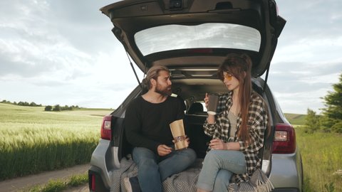 Loving couple sitting in car trunk with coffee and snacks