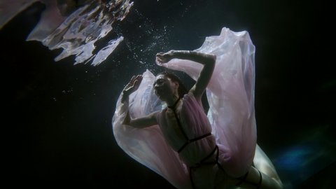 seductive young woman in flowing dress is floating in dark depth of sea, underwater shot