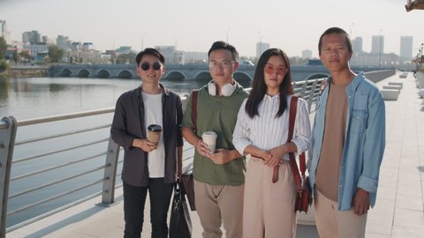 Medium portrait of group of four young Asian friends in smart casualwear posing for camera on river embankment in summer