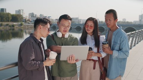 Medium shot of four Asian colleagues using laptop and having conversation standing at river embankment on sunny day