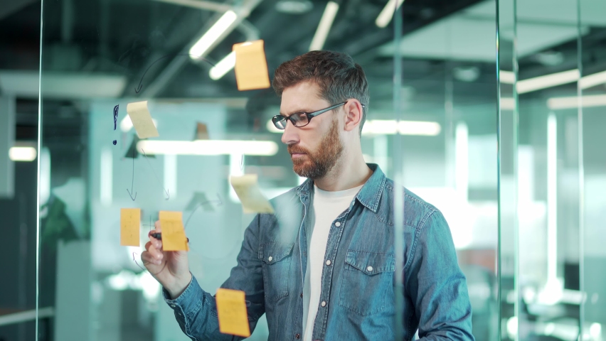 creative young entrepreneur business man in modern office uses post Papers sticky notes to share idea. brainstorming, Bearded thinking male solving strategy Using glass note wall Board. startup posted Royalty-Free Stock Footage #1084623697