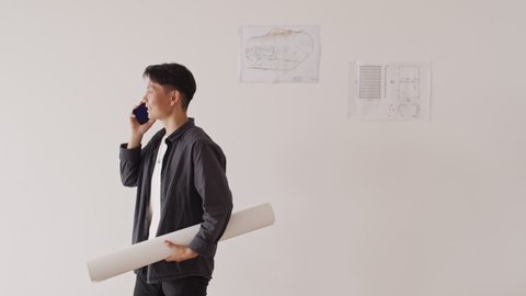 Medium shot of gender-fluid Asian architect with rolled house plan in hands talking on smartphone standing against white wall