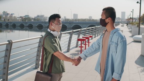 Medium shot of two young Asian men in casualwear and face masks shaking hands standing outdoors at river embankment on summer day