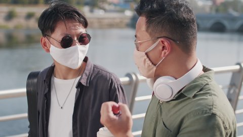 Tracking waist up shot of two Asian friends in face masks having conversation while walking along river embankment on sunny summer day