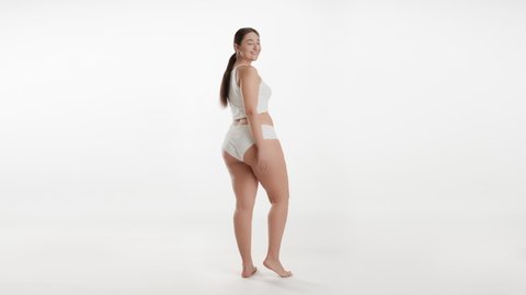 Young Caucasian plus size female model with long dark hair in ponytail in white underwear turns her head to the camera smiling wide and goes away on white background | Skincare concept
