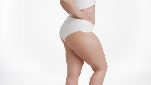 Horizontal medium shot of white-skinned plus size woman in white underwear strokes her thigh and puts her hand on her waistline turning sideways on white background | Unwanted leg hair removal concept