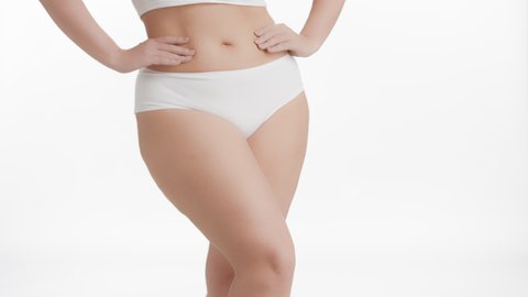 Horizontal medium shot of white-skinned plus size woman in white underwear strokes her thigh and puts her hands on her waistline on white background | After leg shaving concept