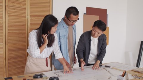 Medium shot of team of Asian architects having discussion while standing by desk and looking at rolled out house plan making measurements