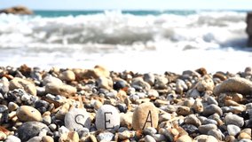 Hand puts stones with letters. Word Sea is written on stones close-up, against background of sea stones, waves. Letters are written on stones sea. Relax, spa, summer vacation sea. Slow motion video.