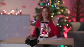 blog about new year decor, young woman with scissors cuts snowflake out of paper sitting at table at home in front of the camera of mobile phone against the background of Christmas tree