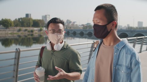 Tracking waist up shot of two young Asian men in face masks chatting during walk along river embankment in summer
