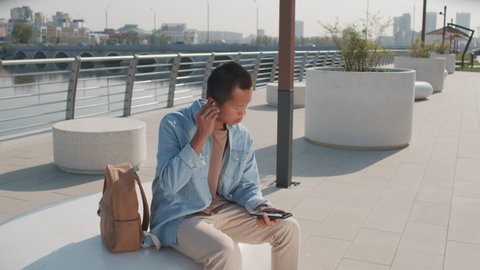 Medium shot of young Asian man in wireless earphones and casualwear texting on smartphone sitting at river embankment on sunny day