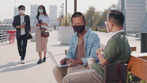Medium shot of two young Asian men in face masks discussing work while sitting on bench at river embankment in summer where two young Asian women strolling by with takeaway coffee