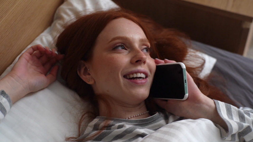 Close-up top view of happy young woman lying on bed and talking mobile phone in evening with friend at home. Cheerful redhead female having pleasant conversation on smartphone, slow motion. Royalty-Free Stock Footage #1084630630